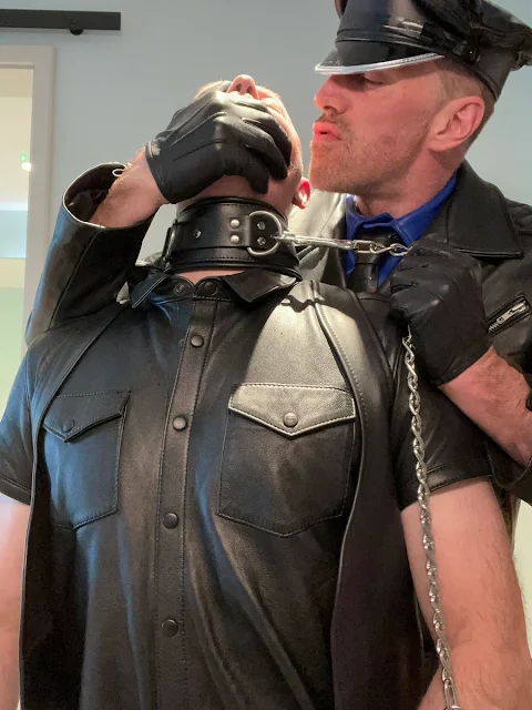 Leathermaster in wear cap holding slave by the collar