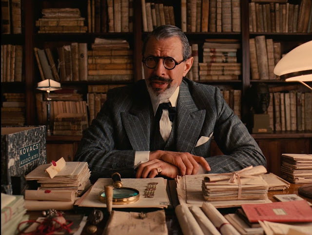 Cena de "O Grande Hotel Budapeste". Mostra um homem sentado a uma mesa. A parede atrás dele está coberta com estantes cheias de livros.