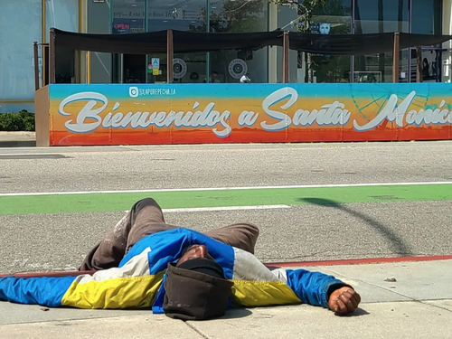 A homeless man lays on the sidewalk across from a sign saying “Welcome to Santa Monica” in Santa Monica, Calif. (Courtesy of The Santa Monica Coalition)