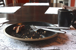 image of a black plate with crumbs from a waffle; a silver fork and knife rest diagonally on the plate; table is dark brown with placemats with a white/black/gray design