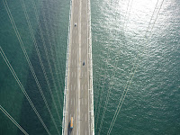 Looking down at the traffic on the road deck of the Akashi Kaikyo Bridge from the top of one of the main towers. The main cables are not visible bu supportieng cables that attach the main cable to the roadway are visible.The ocean is a deep green colour.