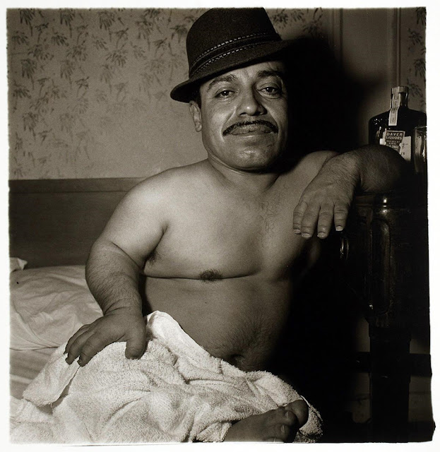 Foto em preto e branco, mostrando um homem anão, em um quarto sentado na cama, sem camisa, com uma toalha na cintura e um chapéu na cabeça. O homem também tem um bigode e apoia o cotovelo esquerdo na cabeceira da cama enquanto sorri olhando para a frente