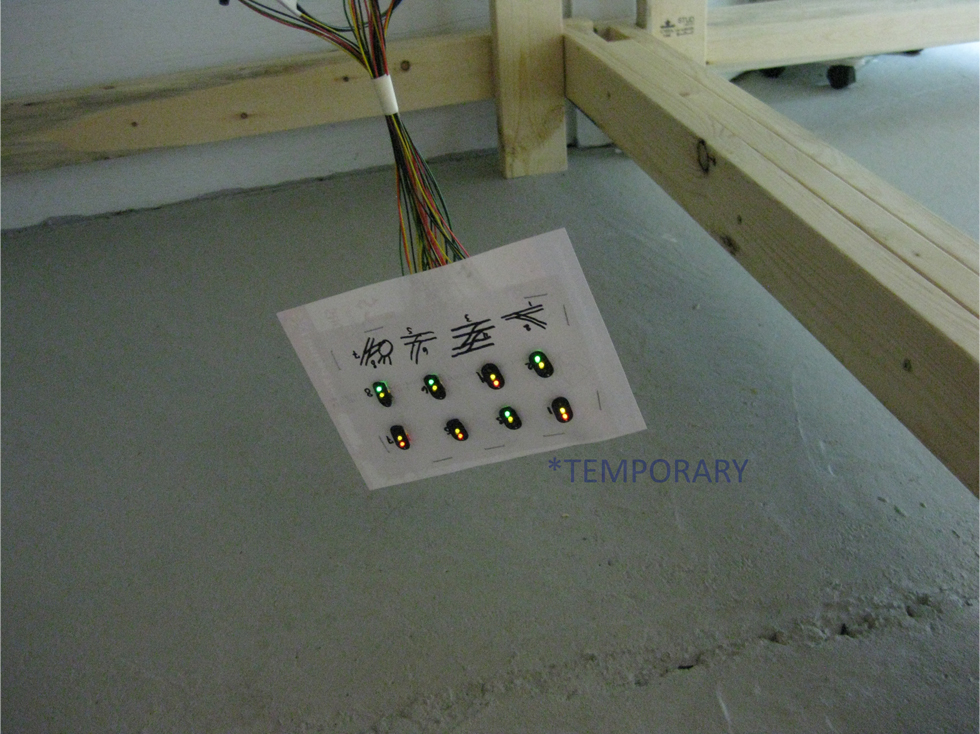 8 trackside signal face plates with LEDs temporarily attached to a sheet of cardstock hanging from under benchwork