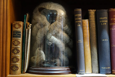 Part of a shelf filled with old books with legal titles and a glass dome with a barrister's wig inside.