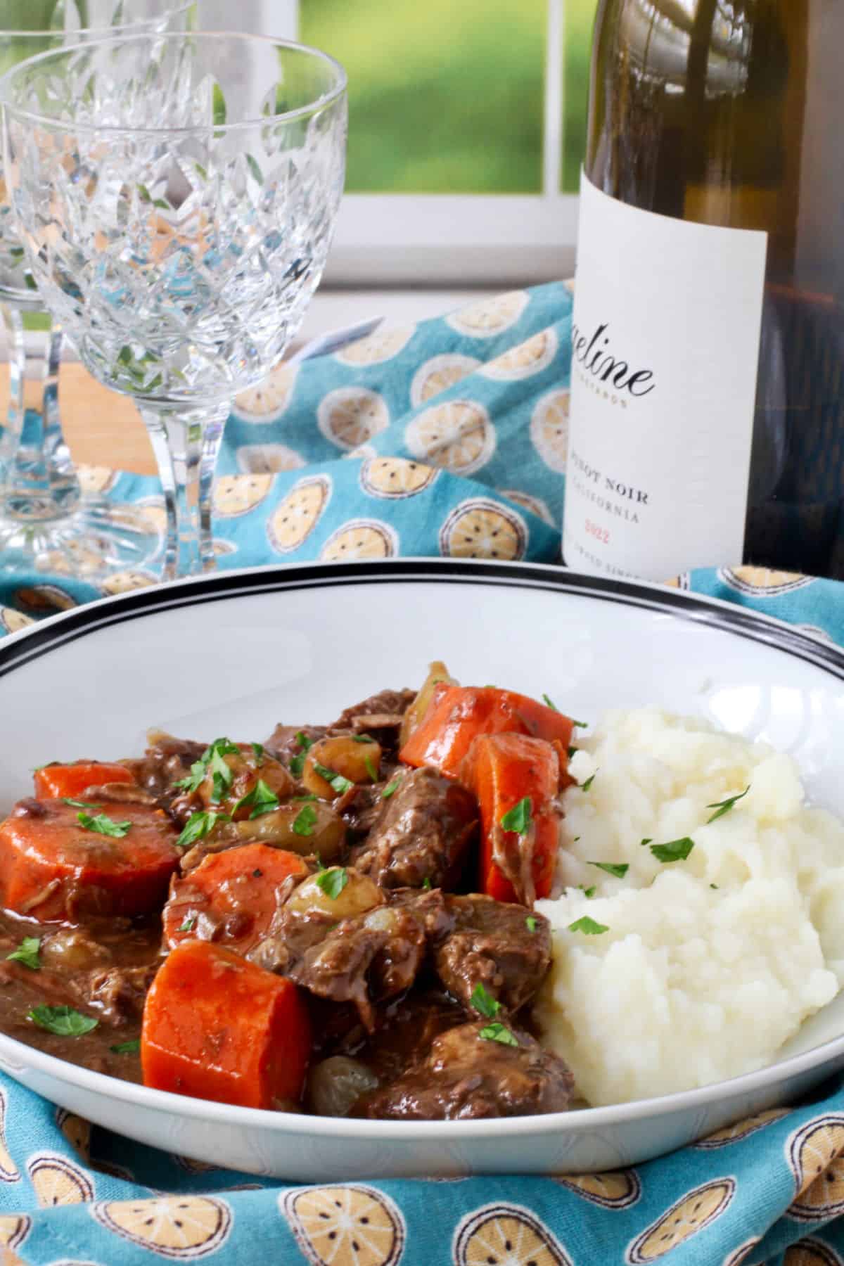 Boeuf Bourguignon in a bowl with wine glasses and wine in the background.