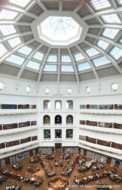 A tall white cupola over a three-storeyed property with a large circular room at its centre with walls lined with bookshelves and people sitting randomly at 22 long wooden desks with several green reading lights.