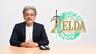 Eiji Aonuma at a desk, with the Tears of the Kingdom logo right next to him and a pair of white Joy-Cons in a grip