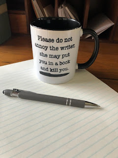 A mug saying "Please do not annoy the writer she may put you in a book and kill you" sitting on top of a note pad next to a pen.