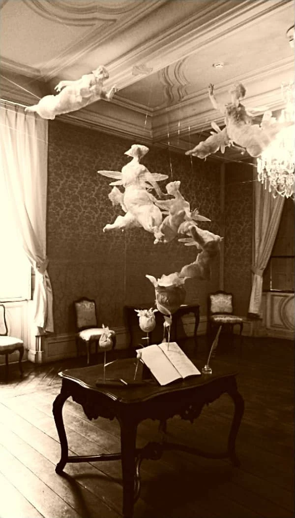historical, high ceiling room contains mobile of all-white paper sculpture angels circling above writing desk on which an open book and paper flowers are placed