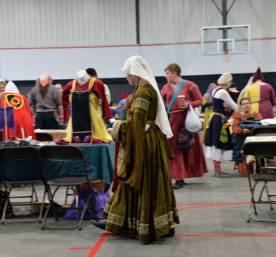 A full-length photo of a white, redheaded woman with pigtail braids falling forward over her shoulders, walking left and looking at the red-and-yellow outfit on the green-draped table to her right. She's wearing a translucent white veil low on her forehead, which falls just past her waist. Her dress is olive-green with sleeves falling to her knees and dense gold embroidered trim of vines and oak leaves bordered by rows of exes, worked on brown silk, and encircling the collar, hem, upper arms, and sleeve hems. Her arms are loosely bent and hands probably clasped, though out of sight. She has a belt in narrow stripes of saffron and maroon. One black, pointed-toed shoe shows as she takes a step. Behind her are more people in bright medieval clothing, standing and talking to each other around tables with items on them.