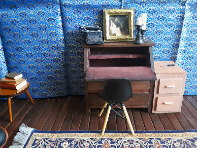 One-tweltfh scale modern miniature dark academia study with a piece of patterned fabric clipped to the wall behind a writing desk and chair