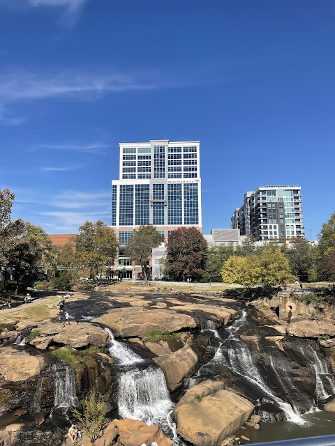 A picture from that scary bridge over the river in Greenville, South Carolina. In front of us are a half dozen separate waterfalls falling into the river below. The city buildings are surrounding the water. The sky is a brilliant blue.