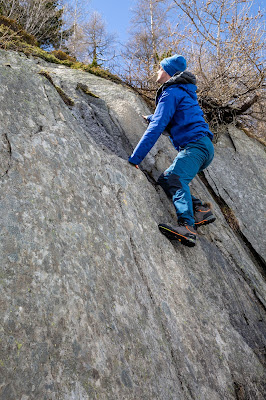 A young white man in a blue rain jacket and the SCARPA Zodiac Plus GTX boots climbs a rock slab.