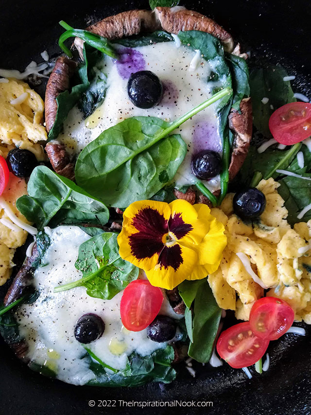 Portobello mushrooms steaks with scrambled eggs, spinach leaves, blueberries, cherry tomatoes and yellow pansies in a skillet.