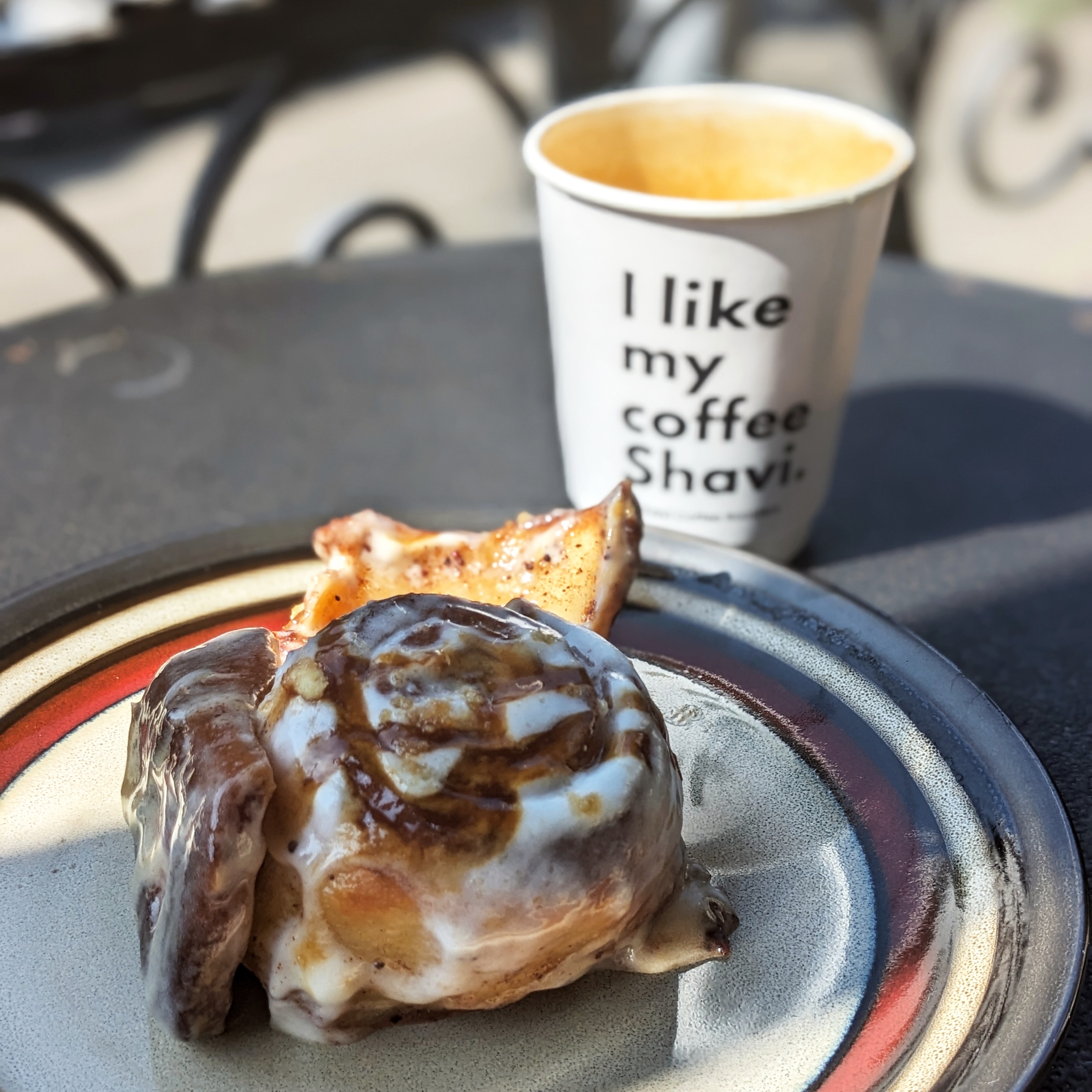 An iced cinnamon bun on a plate with a paper cup of coffee in the background, the coffee and bun are from Shavi Coffee Roasters in Tbilisi, it's one of the best places to go in the city for the best breakfast