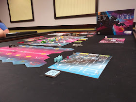 The game Black Angel ready to be played. The main board is seen in the midground, with the chevron-shaped tiles forming the modular space-travel board on which the Black Angel spaceship figure sits. The box sits in the background.