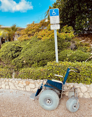 A beach wheelchair with balloon tires is shown sitting atop gravel, parked underneath a "handicapped parking" sign, with shrubbery in the background.