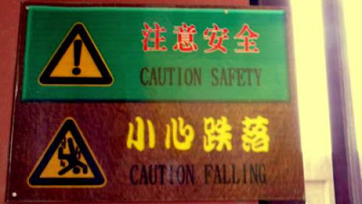 Image of a warning on the wall in the belfry in Dali, Yunnan Province, China. It reads 'Caution' with an image of an exclamation mark in a yellow warning triangle. Underneath it is an image of a person plunging, headfirst, through the air, with the slogan 'Caution, Falling'...