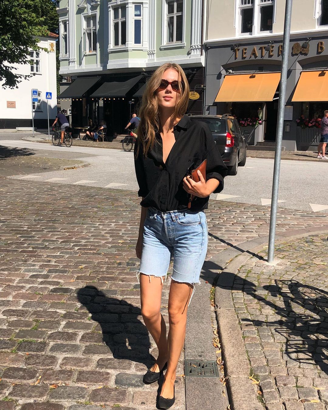 How to Wear Denim Bermuda Shorts — Cecilie Moosgaard Nielsen spring or summer outfit idea with a black button-down shirt, wallet clutch bag, and black ballet flats
