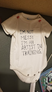 A small white baby onesie with black text that says, "I'm not messy I'm an artist in training."