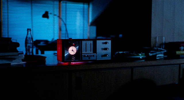 In the bluish light of a darkened room, a rectangular 1960s style clock radio gives off a reddish light.