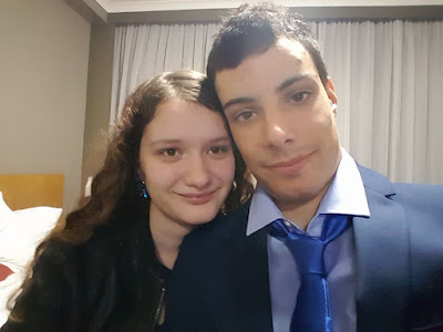 An image of Cy pre-transition and their partner. Cy is white with long brown hair, wearing a leather jacket and blue earings. Their partner has light brown skin and has very short, black hair. He is wearing a dark blue suit and a blue tie