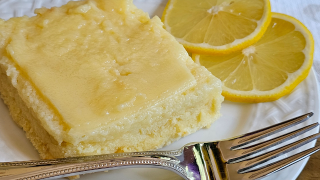 A piece of lemon pudding cake on a white plate with a fork and two slices of fresh lemon.