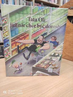 Okładka książki pt. Tata Oli już nie chce być dorosły z rozbrykanym tatą, którym opiekuja sie dzieci. Ksiazka stoi na stoliku. W tle widac regały z ksiazkami.