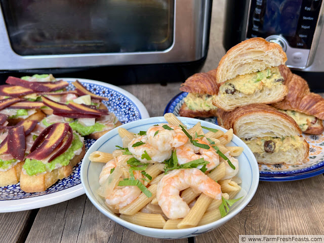 image of an Instant Pot and an air fryer in the background, with a plate of Pea, Artichoke, Carrot & Prosciutto Toasts, Pressure cooker shrimp scampi, and curried chickpea salad sandwiches on croissants in the foreground