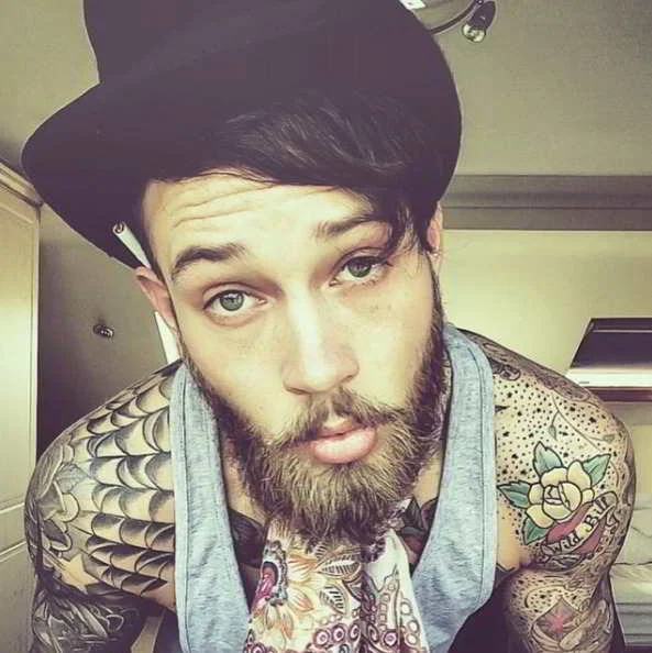 Joven hipster con barba y sombrero lleva tatuaje de tela de araña en el hombro