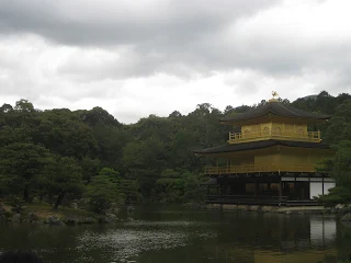 Cloudy sky, blue lake, and large forested area with a 3 story Japanese style palace covered in gold leaf
