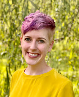 headshot of person with short purple hair, smiling and wearing a yellow shirt with a tree behind them.