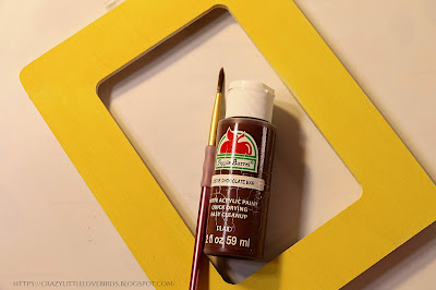 Yellow painted picture frame, smaller paintbrush, and brown paint on a table
