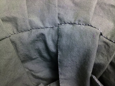 A close-up of a curved, somewhat messily whipstitched seam between matching grey-blue pieces of fabric. Regular pleats fan out along the upper piece of fabric, opening to the right, and a few larger, rougher pleats show on the lower piece of fabric.