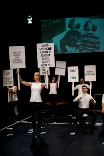 Five people on stage wearing white T-shirts and black pants holding signs with slogans in the air: “I’M DEAF. I DON’T UNDERSTAND A SPEAKING TEACHER”; “I’M NOT ALLOWED TO SIGN AT HOME EITHER”; “LEARNING SPEECH DOESN’T MAKE ME HEARING”; “1880-1981”; “PATERNALISM”; “ADAPTION”). Behind them picture a photograph is projected showing two young boys – hands folded on their back and wearing headphones – from behind.