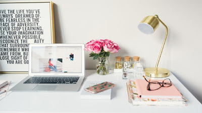 White computer, red roses in a glass vase, mobile phone, folded eye glasses on a pink notepad and a gold lamp holder on top of a white affiliate marketing work station.