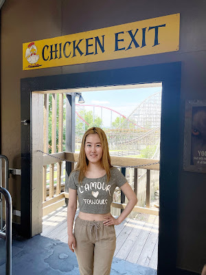 Eunji, an Asian girl with bleached hair wearing brown slacks and a gray shirt, stands in a doorway with a rollercoaster in the background. Above the doorway is a sign that says "Chicken Exit."