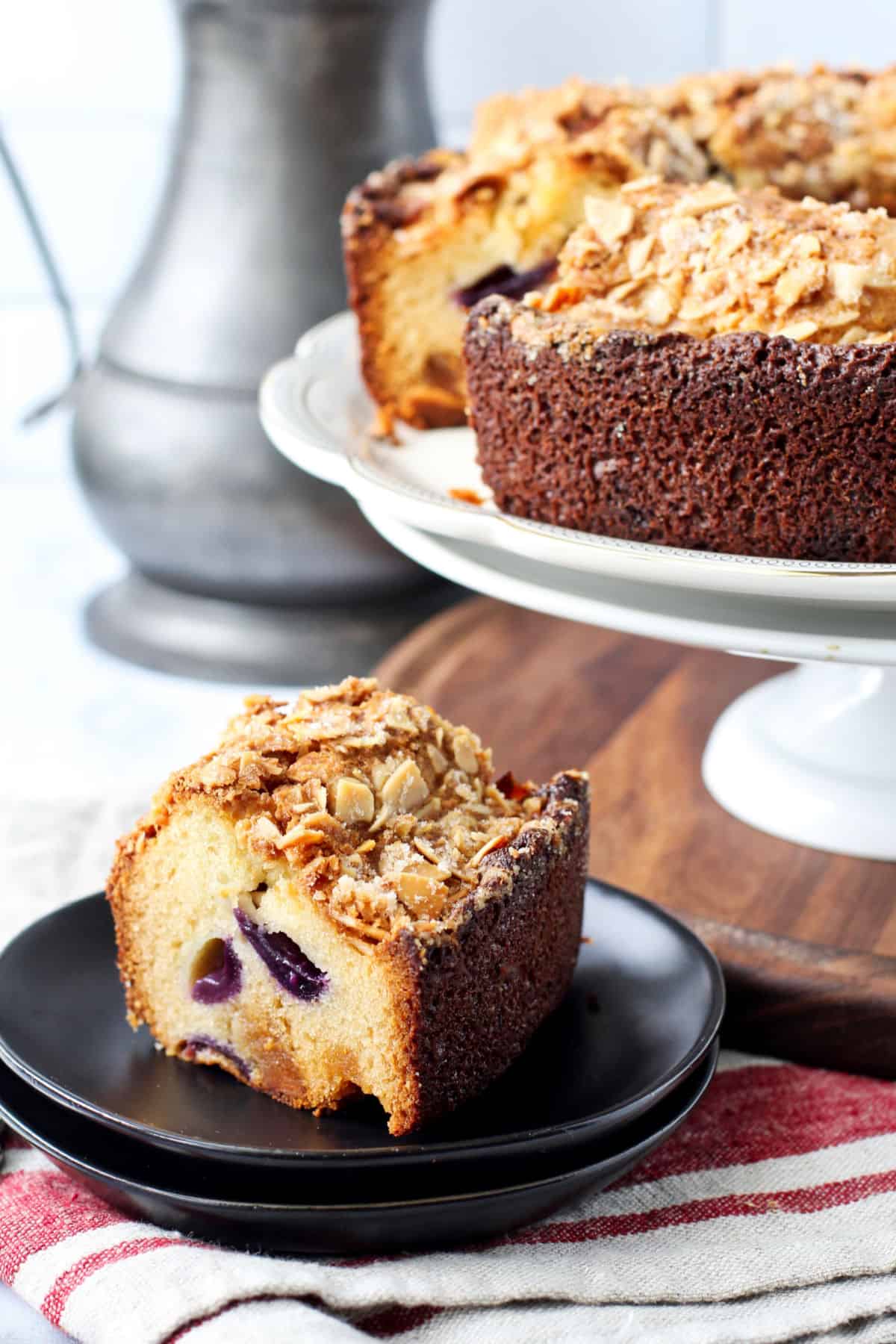 Grape and Almond Paste Cake on a stand with a piece on a black plate in front.