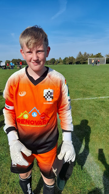 A has sweaty blond hair, is wearing an orange and black goal keepers top, black shorts and white goalie gloves. He has a captain armband on and is smiling