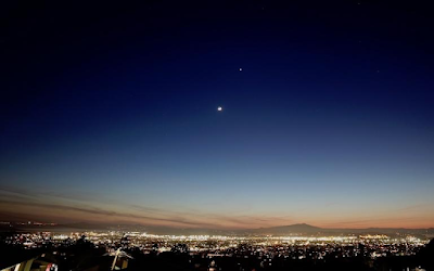 ID: a photo of Venus, the morning star, over a brightly lit city as dawn appears on the horizon. Photo by Jeremy Miller