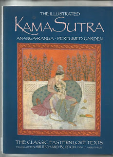 Foto da capa do livro Kama Sutra. A capa é azul marinho. Ao centro, está um casal de amantes em cena ao ar livre. A cena está inserida em uma moldura marrom terracota com arabescos (uma combinação de formas geométricas semelhantes às formas de plantas) em cor de laranja.  Em um terraço, com piso claro, está uma grande almofada ovalada estampada em azul. Atrás, uma mureta clara.  Atrás da mureta, sobre um fundo cor de laranja, despontam longos galhos com pequenas flores azuis.  Os amantes vestem roupas claras, longas e leves: ele, em tons de rosa, ela em tons de verde azulado. Ele usa turbante com fios de pérolas, brincos de argolas e braceletes. Ela usa muitos colares, brincos, anéis e pulseiras. Ambos estão sentados e descalços.  O homem recosta na almofada, a mulher senta de costas para ele e aconchega o corpo entre as pernas masculinas. A mão dele em concha, envolve o seio direito dela.  Ela olha acima, para o amado.  Ela coloca a mão esquerda no pescoço dele e desce a outra mão até a barra do translúcido e feminino véu. Ela cruza a perna direita em formato do número 4.  Ao lado direito, duas bandejas com alimentos e um pequeno pote bojudo completam a ambientação.