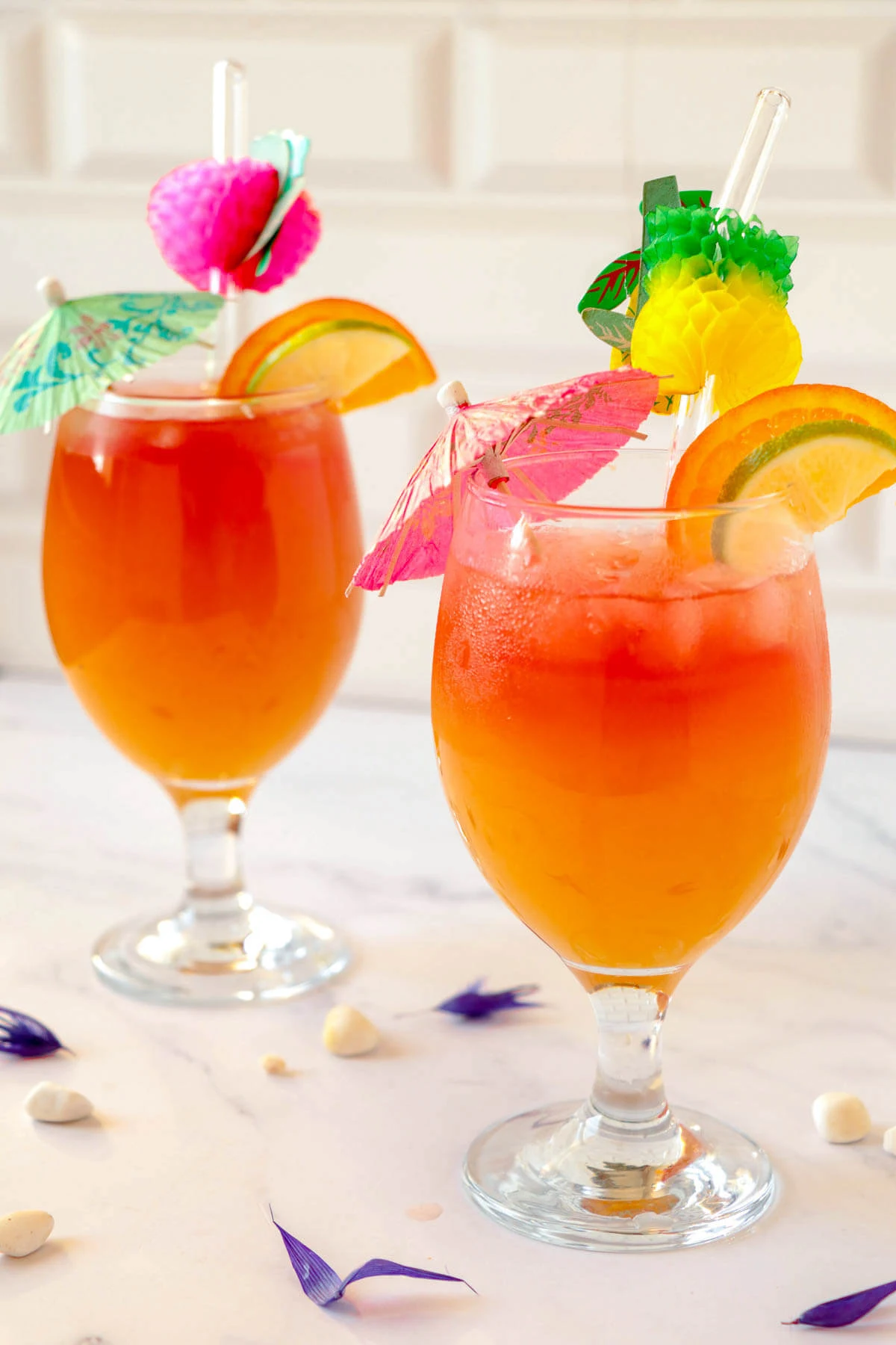 Two full glasses of Caribbean rum punch with slices and umbrellas over some ice.