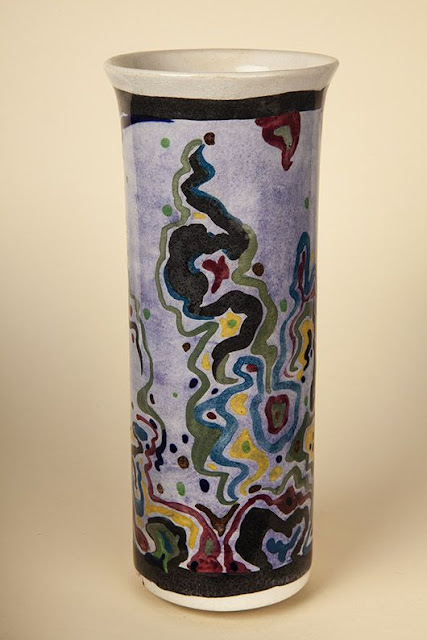 A tall, thin cylindrical vase, made and hand-painted by Albert Wainwright. The painting style is abstract, with purples, reds, yellows, greens and black swirls painted onto the white base.