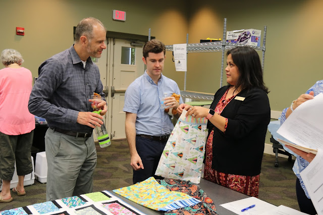 A woman is showing two men a sheet of colorful fabric. They are listening to her intently as she talks. They are in a library.