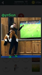Мужчина с игрушкой около телевизора, по которому показывают футбол