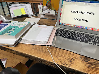 A messy desk. There's papers and notebooks, some post its, and the laptop screen which has a document called Leeza McAuliffe Book 2 on it.