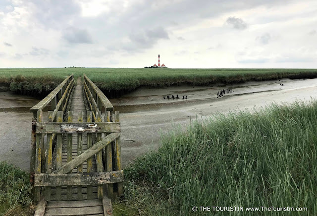 Wooden bridge over a creek in the wadden sea leading to a red and white lighthouse.