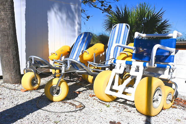 Three, manual beach wheelchairs are parked next to each other. The first two look like lounge chairs, and have one large wheel in front, plus two large wheels in the back. The canvas of the seats is blue- and white-striped. On the right is a beach wheelchair with four tires. The two in the front are larger than the two in the back. There are white armrests, and a blue canvas that acts as the seat, as well as the seat back.