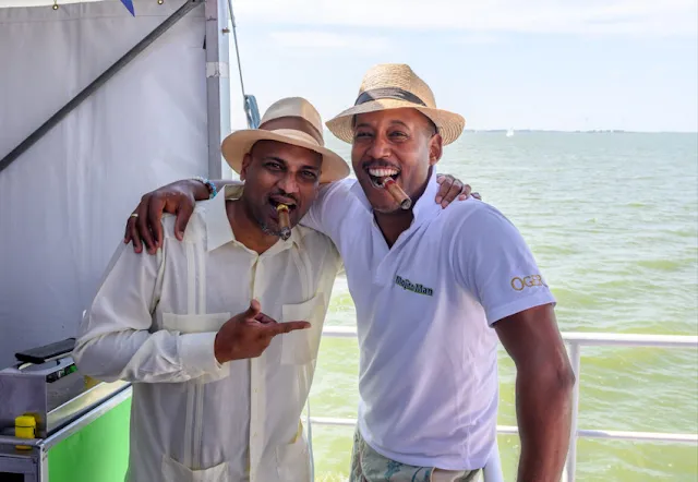 Two black fellows wearing Panama hats and smoking cigars and very happy from the waist up on a boat deck