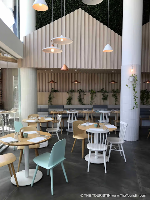 Cafe with pastel green and white chairs on wooden tables on concrete flooring. White lamps. hanging from a high ceiling